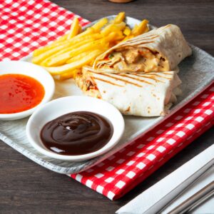 Close-up of a tasty fast food meal including wraps, french fries, and dipping sauces on a checkered cloth.