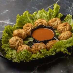 Crispy fried veg momos arranged with spicy sauce on a black platter, garnished with fresh lettuce.