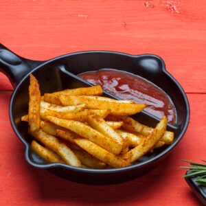 Crispy seasoned French fries with ketchup in a skillet on a red wooden table.